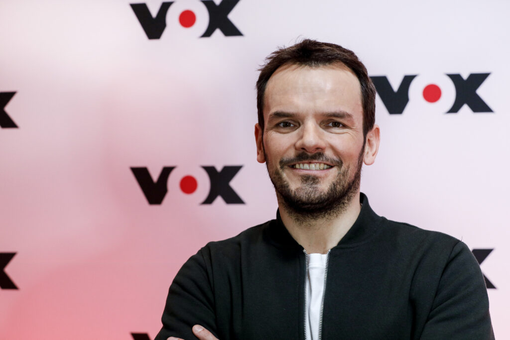 Steffen Henssler steht vor VOX-Werbewand und lächelt in die Kamera