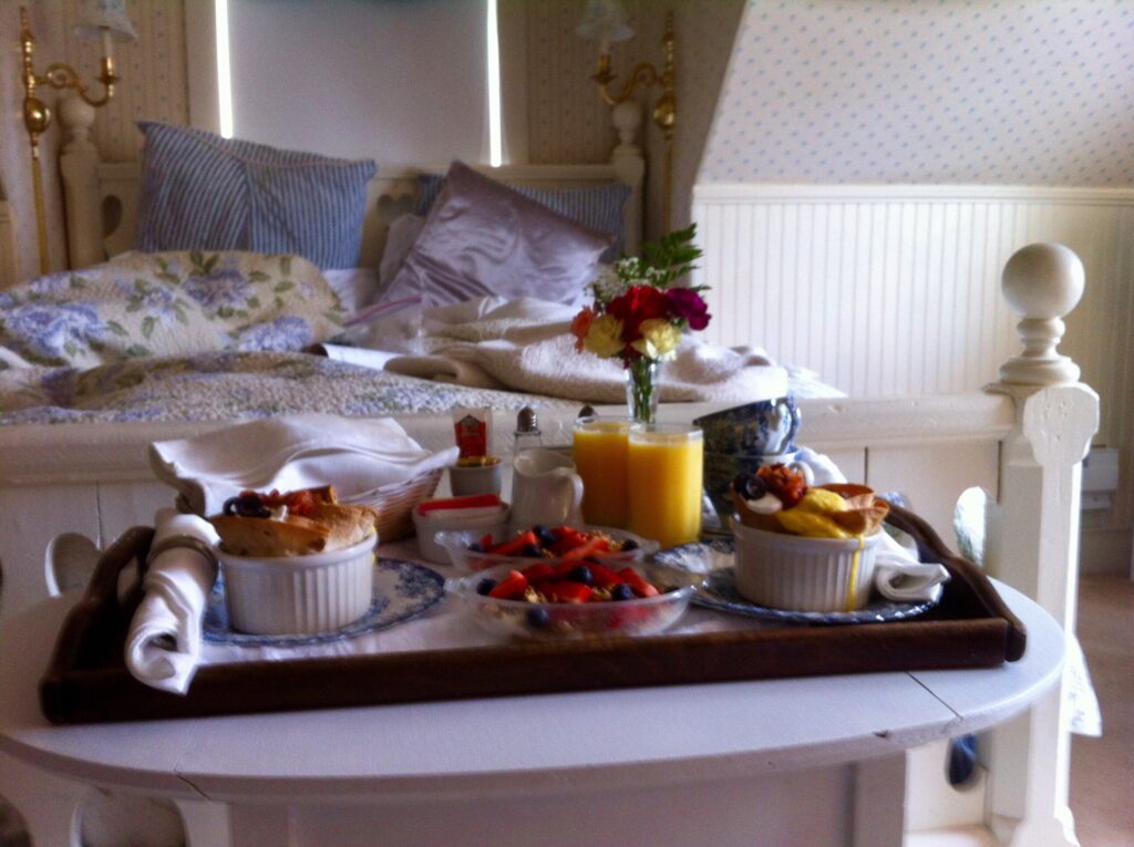 Schlafzimmer mit Tablet davor, auf dem zwei Gläser Organsaft, Blumen, Früchte sowie zwei warme Speisen stehen