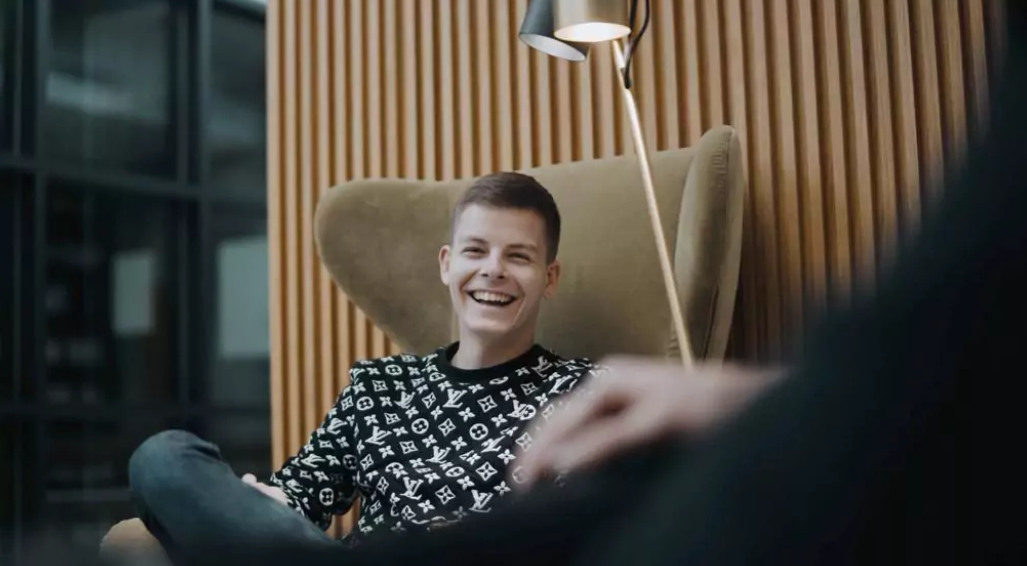 Jürgen Schroll sitzt mit schwarz-weiß kariertem Pullover in einem Sessel und lacht