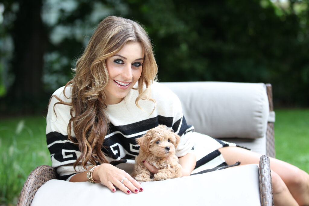 Verena Kerth sitzt mit einem kleinen Hund in der Hand auf einem Gartensofa  und lächelt in die Kamera