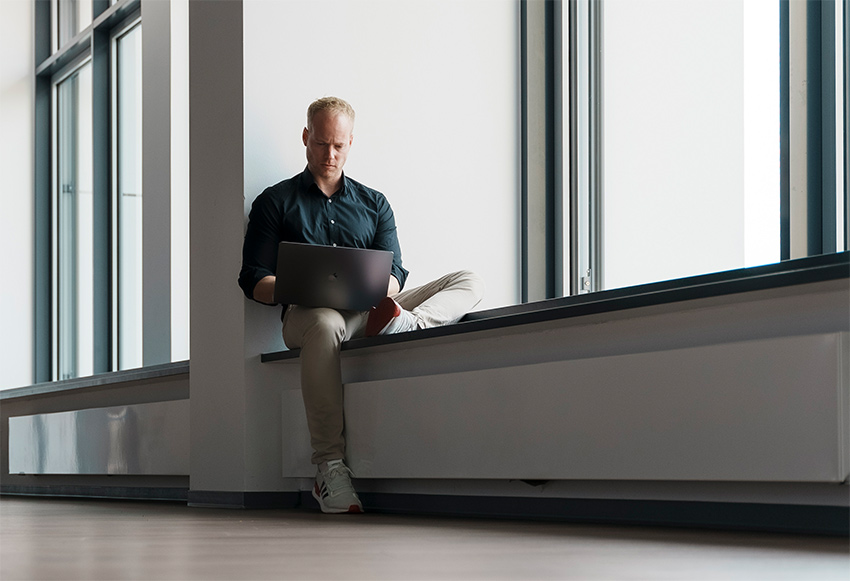 Simon Feller sitzt mit dunklem Hemnd und heller Jeans mit seinem PC auf einer Fensterbank einer langen Fensterfront