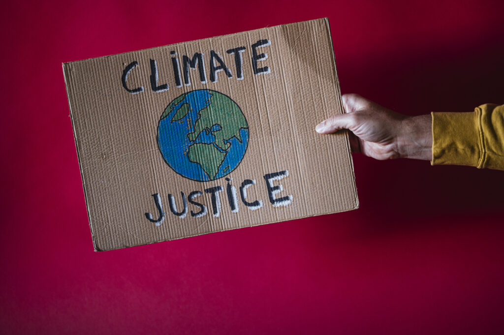 Roter Hintergrund vor dem eine Hand ein Plakat aus Karton mit der Aufschrift CLIMATE JUSTICE hält. In der Mitte ist die Erdkugel abgebildet