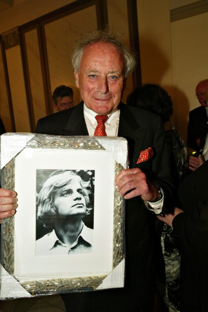 Reinhold Würth hält ein Bild mit silbernen Rahmen und einem jungen Mann in seinen Händen und lacht in die Kamera