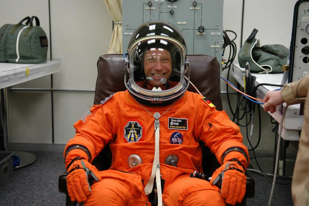 Thomas Reiter sitzt in einem orangen Raumanzug und schwarzem Helm in einem Sessel und lächelt in die Kamera