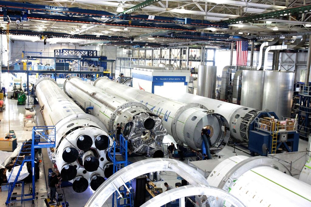 Ein Montagewerk für Elektroautos neben einem Raketenstartturm symbolisiert die mögliche Fusion der Unternehmen von Elon Musk – Foto: SpaceX-Imagery / Pixabay